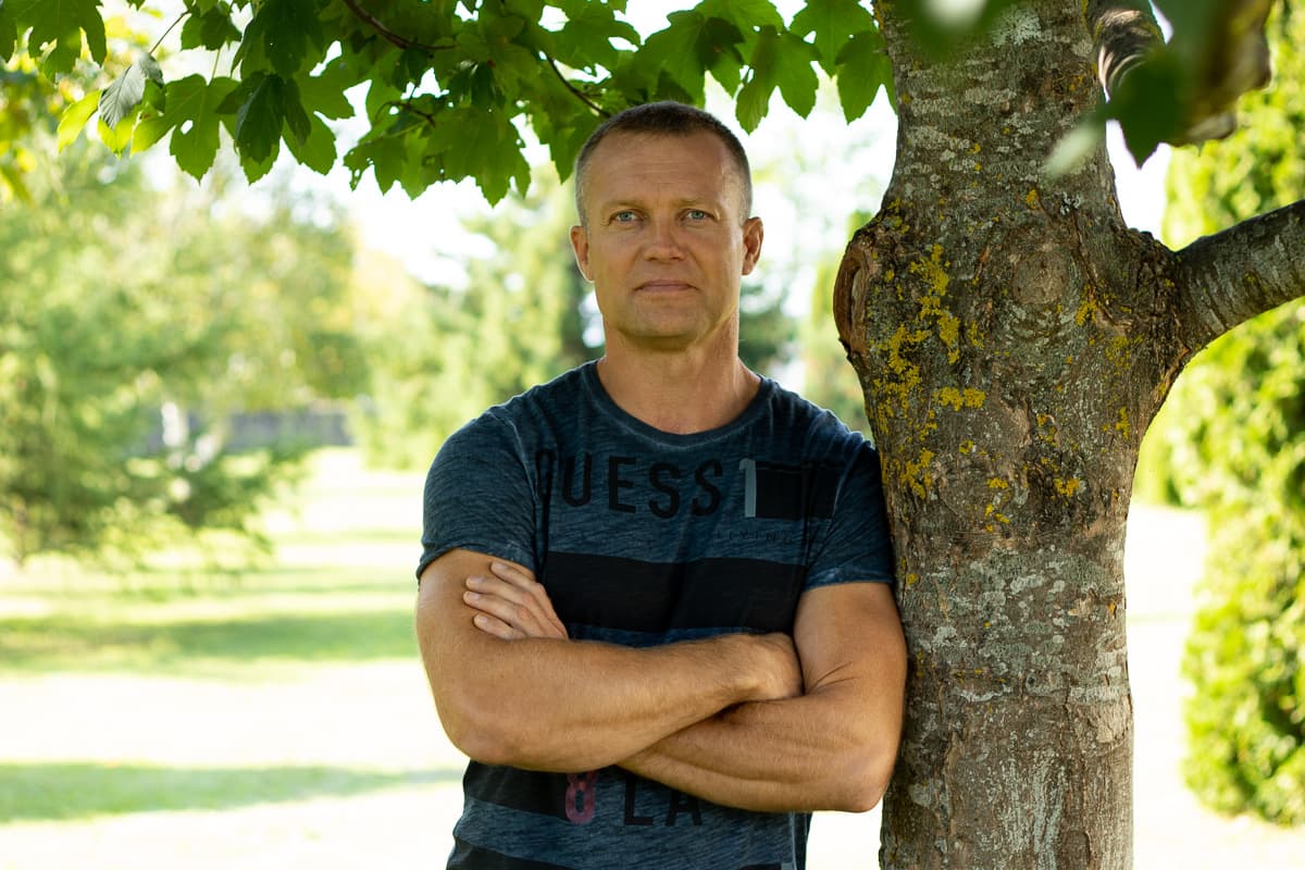 Man with crossed arms leaning against a tree trunk in a sunny, green park.