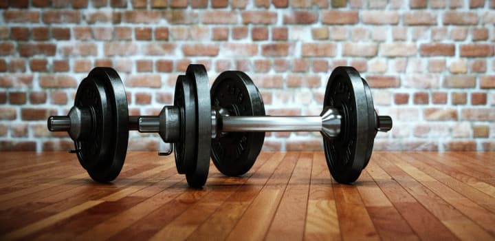 Two heavy black dumbbells resting on a wooden floor against a rustic brick wall.